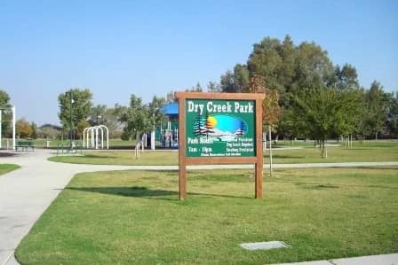 The Dry Creek Park sign on a sunny green lawn in Clovis, California, with a playground behind it